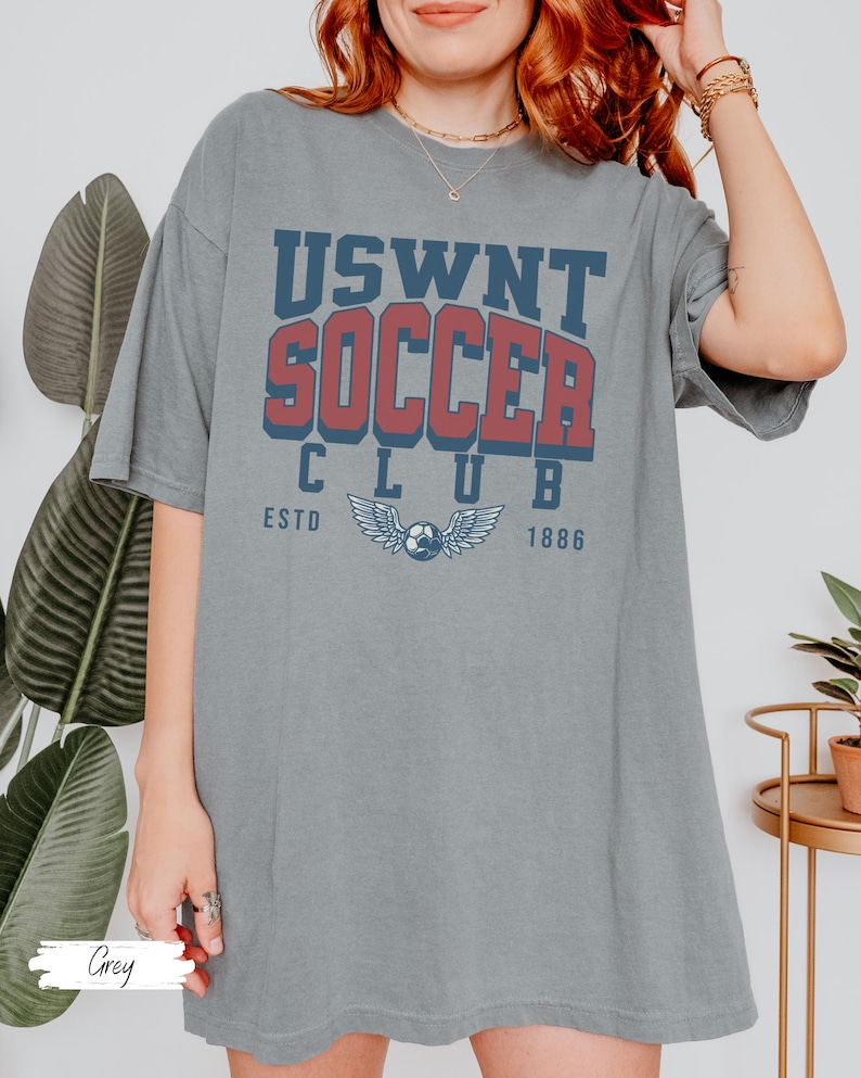 Puede incluir: Una camiseta gris de gran tama&ntilde;o con el texto "USWNT SOCCER CLUB ESTD 1886" impreso en la parte delantera. El texto est&aacute; en una fuente de estilo vintage con un esquema de color rojo y azul. La camiseta presenta un gr&aacute;fico de un bal&oacute;n de f&uacute;tbol con alas.