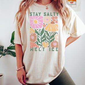 Op de afbeelding: Cr&egrave;mekleurig T-shirt met een bloemenontwerp en de tekst "STAY SALTY MELT ICE". Het ontwerp bevat roze, gele en oranje bloemen met groene bladeren. Het shirt heeft een casual stijl.