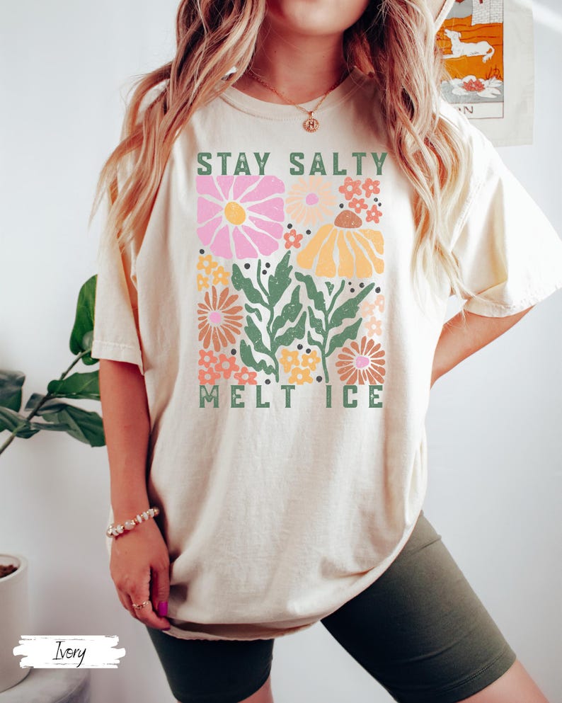 Op de afbeelding: Cr&egrave;mekleurig T-shirt met een bloemenontwerp en de tekst "STAY SALTY MELT ICE". Het ontwerp bevat roze, gele en oranje bloemen met groene bladeren. Het shirt heeft een casual stijl.