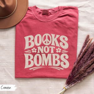 May include: Crimson-colored t-shirt with the text "BOOKS NOT BOMBS" in a retro font. The design includes a peace sign and floral accents. The shirt is a crew neck style.
