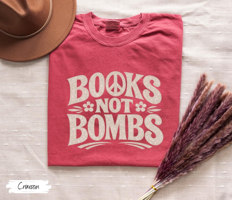 May include: Crimson-colored t-shirt with the text "BOOKS NOT BOMBS" in a retro font. The design includes a peace sign and floral accents. The shirt is a crew neck style.