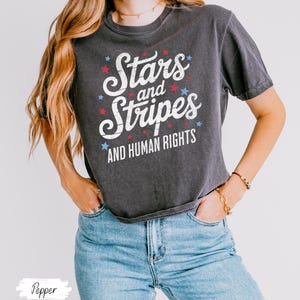 Puede incluir: Camiseta gris oscuro con el texto "Stars and Stripes AND HUMAN RIGHTS" en blanco, rojo y azul. La camiseta tiene cuello redondo y mangas cortas. La modelo lleva vaqueros azules.