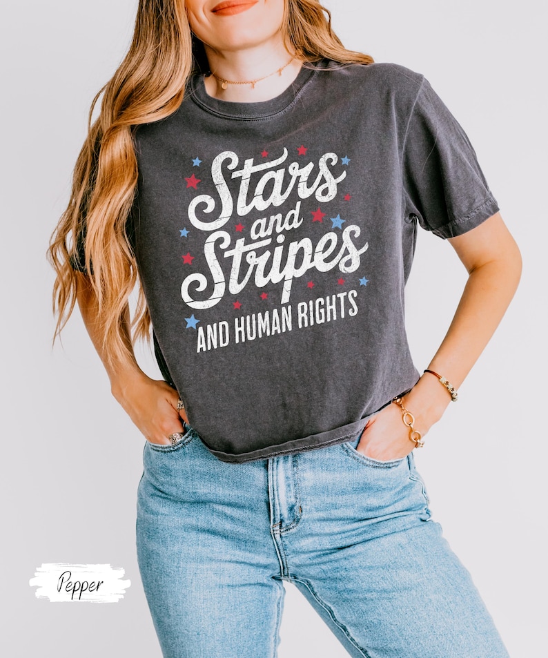 Puede incluir: Camiseta gris oscuro con el texto "Stars and Stripes AND HUMAN RIGHTS" en blanco, rojo y azul. La camiseta tiene cuello redondo y mangas cortas. La modelo lleva vaqueros azules.