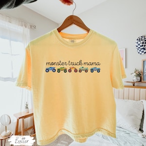 May include: A yellow t-shirt with the text "monster truck mama" above a row of colorful monster truck illustrations. The shirt is hanging on a wooden hanger.