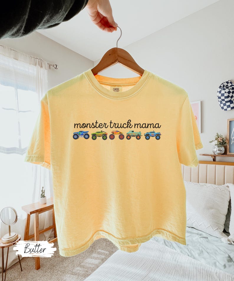 May include: A yellow t-shirt with the text "monster truck mama" above a row of colorful monster truck illustrations. The shirt is hanging on a wooden hanger.