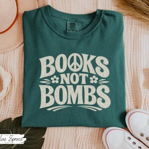 May include: Teal-colored t-shirt with the words "BOOKS NOT BOMBS" in a retro font. The design includes a peace sign and floral accents. The shirt is folded on a light-colored surface, with white sneakers visible.