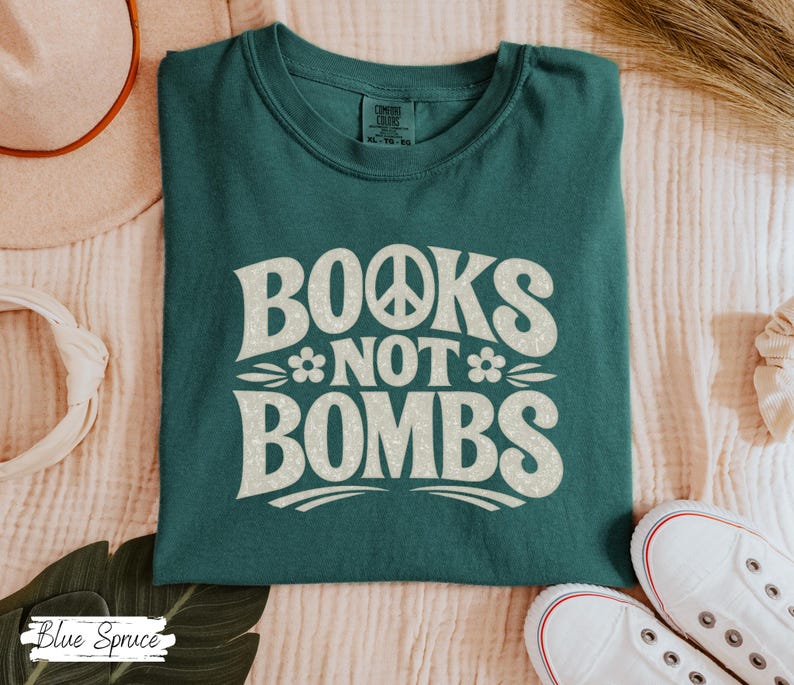 May include: Teal-colored t-shirt with the words "BOOKS NOT BOMBS" in a retro font. The design includes a peace sign and floral accents. The shirt is folded on a light-colored surface, with white sneakers visible.