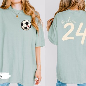 Op de afbeelding: Lichtgroen T-shirt met een voetbalafbeelding en een rood hart op de voorkant. De achterkant van het shirt toont de naam "Pulisic" en het nummer "24" in cr&egrave;me. Het shirt heeft een casual stijl.