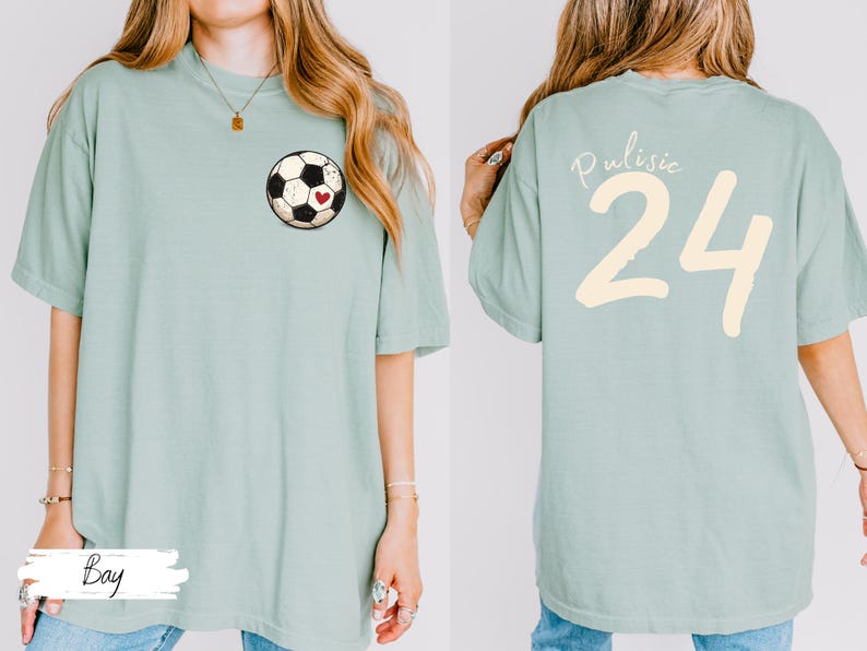 Op de afbeelding: Lichtgroen T-shirt met een voetbalafbeelding en een rood hart op de voorkant. De achterkant van het shirt toont de naam "Pulisic" en het nummer "24" in cr&egrave;me. Het shirt heeft een casual stijl.