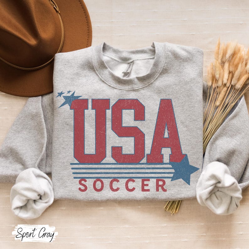 May include: A gray sweatshirt with a red, white, and blue graphic that says "USA SOCCER" with stars on either side.