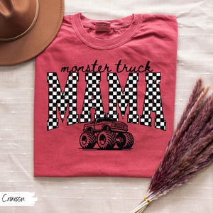 Op de afbeelding: Rood T-shirt met de tekst "monster truck MAMA" in zwart-witte geblokte letters. Een zwarte monster truck afbeelding staat onder de tekst. Het shirt heeft een ronde hals.
