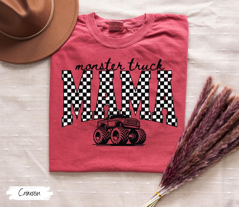 Op de afbeelding: Rood T-shirt met de tekst "monster truck MAMA" in zwart-witte geblokte letters. Een zwarte monster truck afbeelding staat onder de tekst. Het shirt heeft een ronde hals.