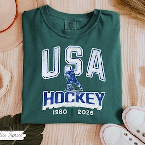 May include: A dark teal t-shirt with the text "USA HOCKEY" in white and blue, featuring a hockey player graphic. The shirt includes the years 1980 and 2026. The shirt is folded and placed on a light-colored surface.