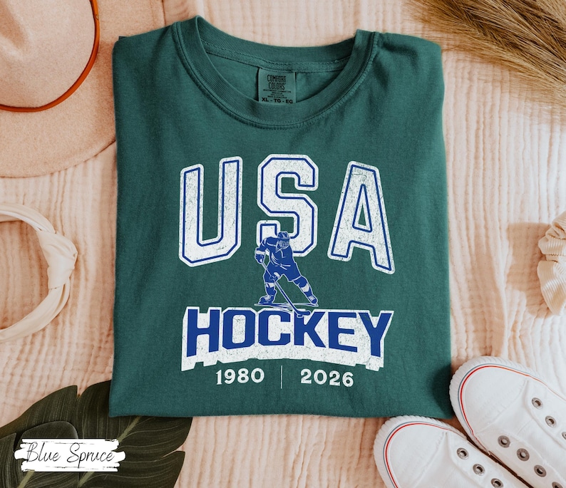 May include: A dark teal t-shirt with the text "USA HOCKEY" in white and blue, featuring a hockey player graphic. The shirt includes the years 1980 and 2026. The shirt is folded and placed on a light-colored surface.