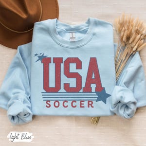 May include: Light blue sweatshirt with a vintage-style graphic of "USA SOCCER" in red and blue.