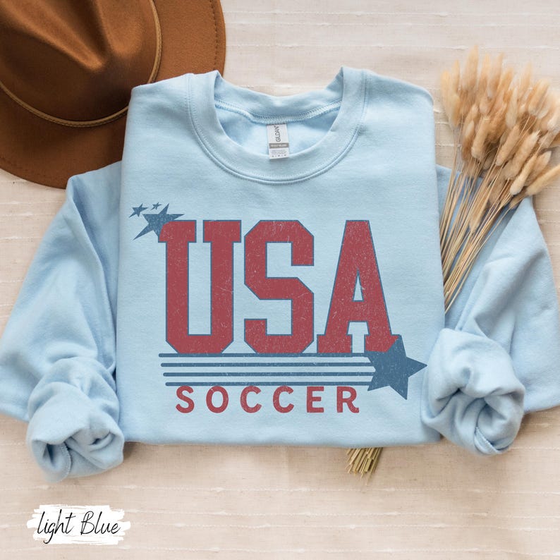 May include: Light blue sweatshirt with a vintage-style graphic of "USA SOCCER" in red and blue.