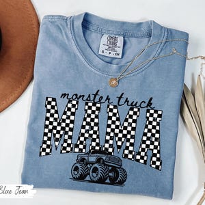 Op de afbeelding: Een blauw Comfort Colors T-shirt met de woorden "monster truck MAMA" in zwart-wit geblokte letters. Een afbeelding van een monstertruck staat onder de tekst. Een gouden ketting is over het shirt gedrapeerd.