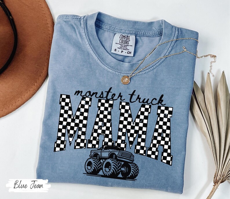 Op de afbeelding: Een blauw Comfort Colors T-shirt met de woorden "monster truck MAMA" in zwart-wit geblokte letters. Een afbeelding van een monstertruck staat onder de tekst. Een gouden ketting is over het shirt gedrapeerd.