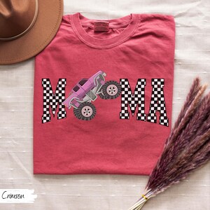 May include: Crimson-colored t-shirt with the word "MAMA" in a checkered pattern, featuring a pink monster truck graphic. The shirt is a crew neck and has a relaxed fit.