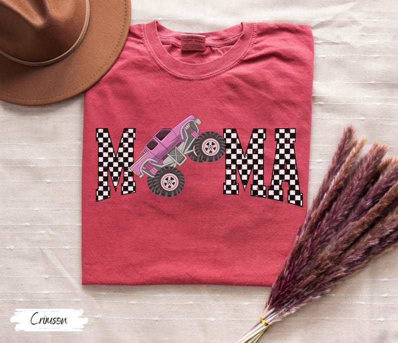 May include: Crimson-colored t-shirt with the word "MAMA" in a checkered pattern, featuring a pink monster truck graphic. The shirt is a crew neck and has a relaxed fit.