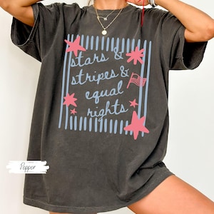 Puede incluir: Camiseta gris oscuro con el texto "stars & stripes & equal rights" en un dise&ntilde;o azul claro y rosa. El dise&ntilde;o incluye estrellas y una peque&ntilde;a bandera americana. La camiseta es de corte holgado.