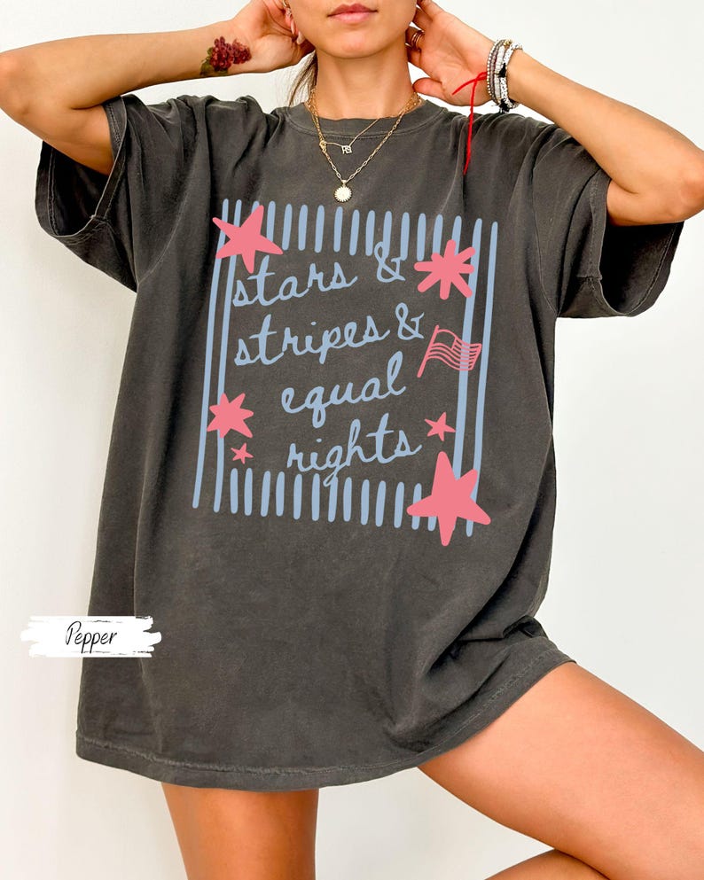 Puede incluir: Camiseta gris oscuro con el texto "stars & stripes & equal rights" en un dise&ntilde;o azul claro y rosa. El dise&ntilde;o incluye estrellas y una peque&ntilde;a bandera americana. La camiseta es de corte holgado.