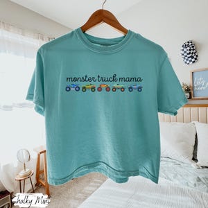 May include: A teal-colored t-shirt with the text "monster truck mama" above a row of colorful monster truck illustrations. The shirt is hanging on a wooden hanger.