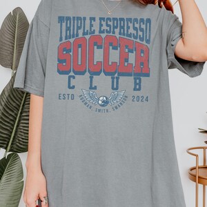 Op de afbeelding: Een grijs oversized T-shirt met de tekst "Triple Espresso Soccer Club" in rood en blauwe letters. De tekst "Estd. 2024" staat onder de clubnaam. Een voetbal met vleugels bevindt zich in het midden van het shirt.