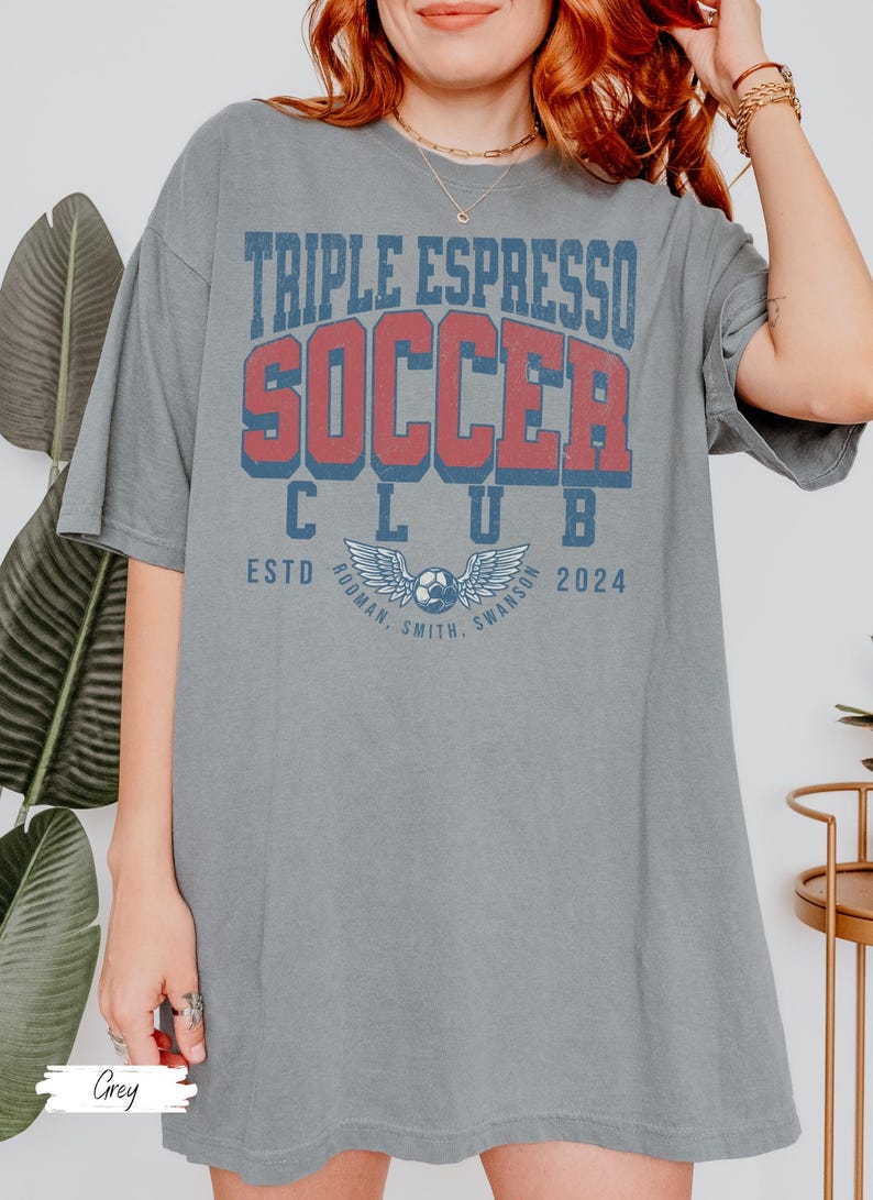Op de afbeelding: Een grijs oversized T-shirt met de tekst "Triple Espresso Soccer Club" in rood en blauwe letters. De tekst "Estd. 2024" staat onder de clubnaam. Een voetbal met vleugels bevindt zich in het midden van het shirt.