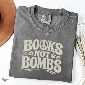 May include: A gray t-shirt with the text "BOOKS NOT BOMBS" in a retro font, featuring a peace sign and flower. A gold necklace with a pendant is draped across the shirt. The shirt is folded, with a brown hat visible in the background.