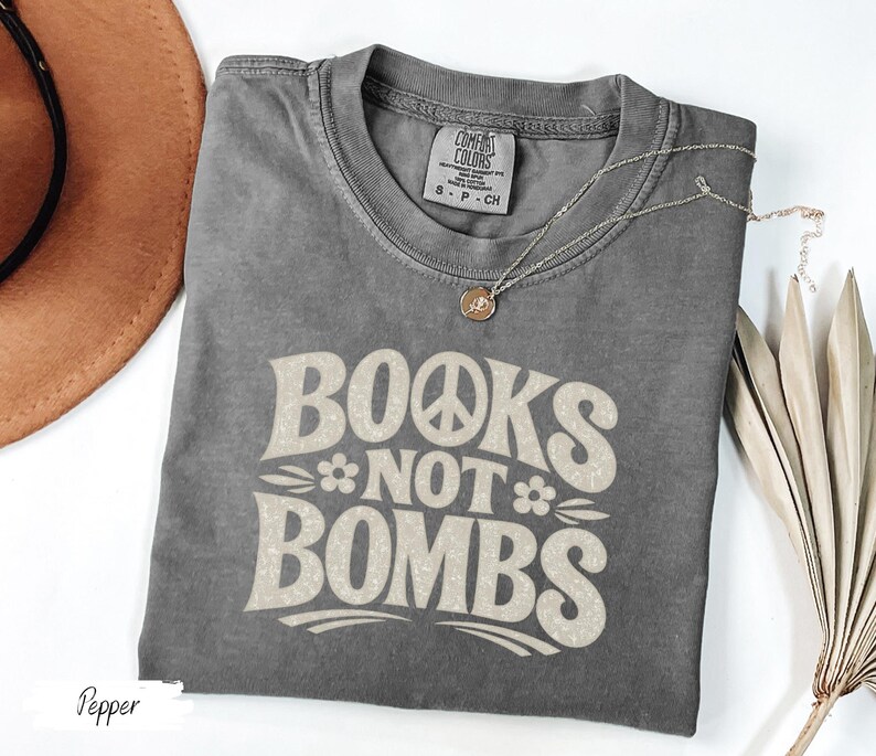 May include: A gray t-shirt with the text "BOOKS NOT BOMBS" in a retro font, featuring a peace sign and flower. A gold necklace with a pendant is draped across the shirt. The shirt is folded, with a brown hat visible in the background.