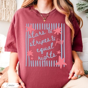 Puede incluir: Camiseta color carmes&iacute; con el texto "Stars & Stripes & Equal Rights" en azul claro. El dise&ntilde;o incluye estrellas, rayas y una peque&ntilde;a bandera americana. La camiseta es de estilo casual y c&oacute;modo.