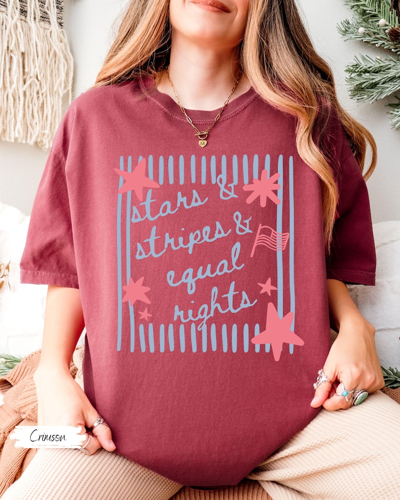 Puede incluir: Camiseta color carmes&iacute; con el texto "Stars & Stripes & Equal Rights" en azul claro. El dise&ntilde;o incluye estrellas, rayas y una peque&ntilde;a bandera americana. La camiseta es de estilo casual y c&oacute;modo.