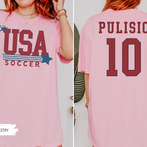 May include: Pink t-shirt with "USA SOCCER" text in red and blue, featuring stars. The back of the shirt displays "PULISIC" and the number "10" in red with a navy blue outline. A casual, sporty design.
