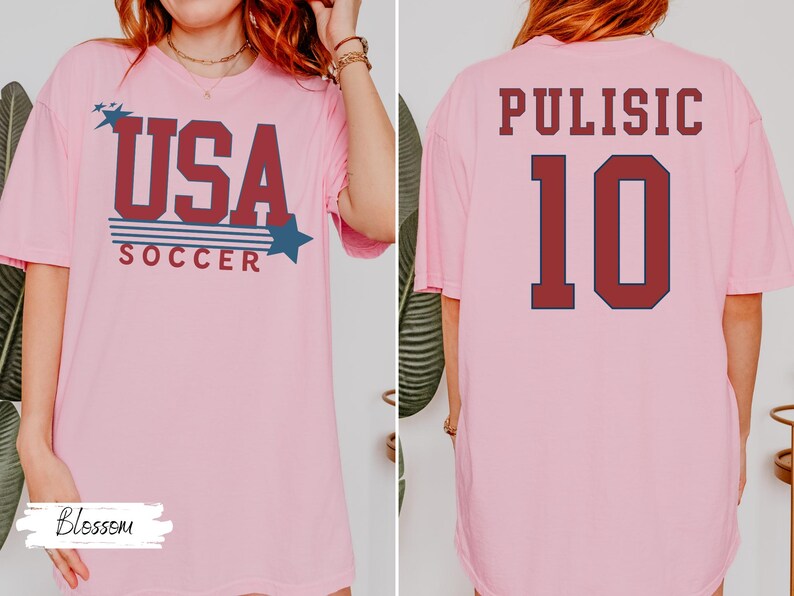 May include: Pink t-shirt with "USA SOCCER" text in red and blue, featuring stars. The back of the shirt displays "PULISIC" and the number "10" in red with a navy blue outline. A casual, sporty design.