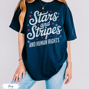 Puede incluir: Camiseta azul marino con el texto "Stars and Stripes AND HUMAN RIGHTS" en blanco, rojo y azul. El dise&ntilde;o incluye estrellas. La camiseta es una prenda informal de manga corta.