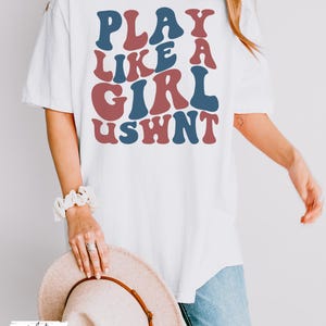 May include: White oversized t-shirt with the text "PLAY LIKE A GIRL USWNT" in blue and red retro font.