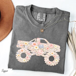 Op de afbeelding: Een grijs Comfort Colors T-shirt met een bloemen monster truck ontwerp. De truck is versierd met roze, witte en blauwe bloemen. Een gouden ketting met een hanger ligt op het shirt. Het woord "Pepper" staat linksonder.