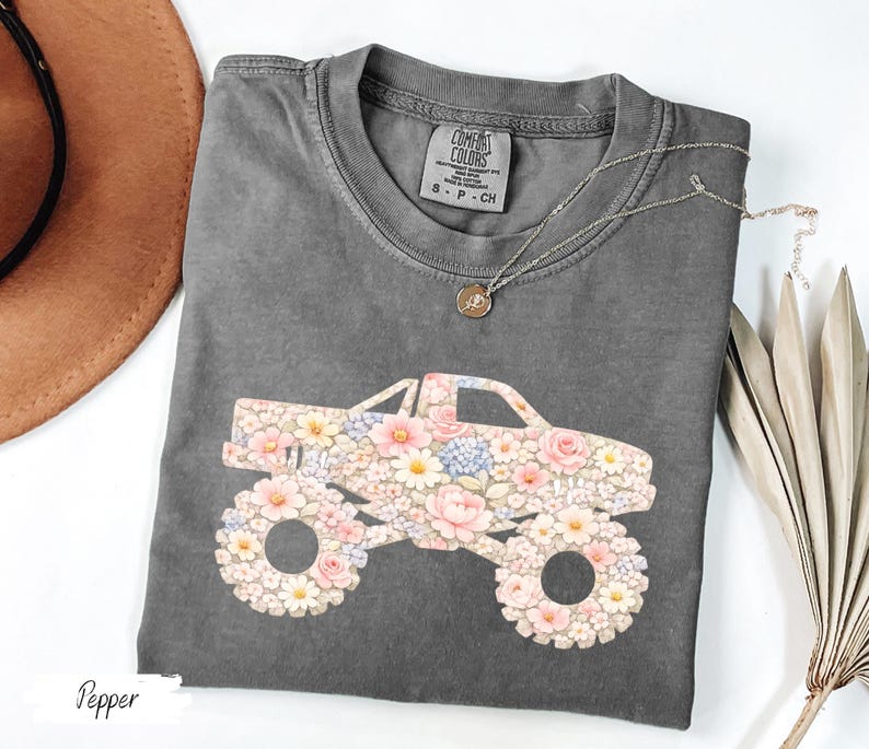 Op de afbeelding: Een grijs Comfort Colors T-shirt met een bloemen monster truck ontwerp. De truck is versierd met roze, witte en blauwe bloemen. Een gouden ketting met een hanger ligt op het shirt. Het woord "Pepper" staat linksonder.