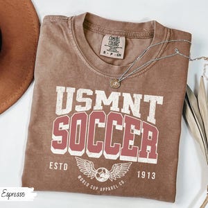 Może przedstawiać: Koszulka w kolorze espresso z napisem "USMNT SOCCER" w białych i czerwonych literach. Koszulka posiada piłkę nożną ze skrzydłami oraz napis "ESTD 1913" i "WORLD CUP APPAREL CO." Srebrny naszyjnik jest zarzucony na koszulkę.