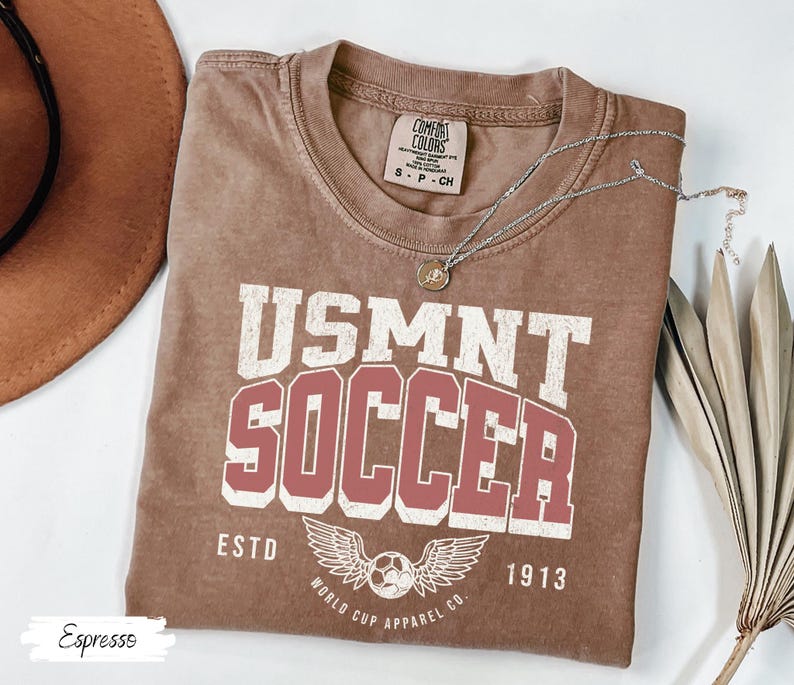 Może przedstawiać: Koszulka w kolorze espresso z napisem "USMNT SOCCER" w białych i czerwonych literach. Koszulka posiada piłkę nożną ze skrzydłami oraz napis "ESTD 1913" i "WORLD CUP APPAREL CO." Srebrny naszyjnik jest zarzucony na koszulkę.