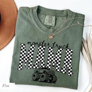 Op de afbeelding: Mosgroen t-shirt met de tekst "monster truck MAMA" in een zwart-wit geblokt patroon. Een monster truck-afbeelding staat onder de tekst. Het shirt is van het merk Comfort Colors.