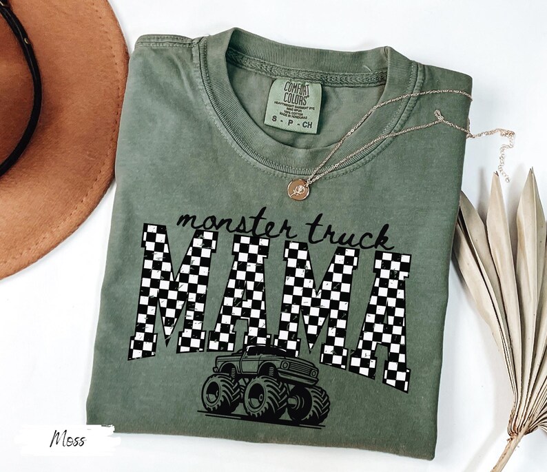 Op de afbeelding: Mosgroen t-shirt met de tekst "monster truck MAMA" in een zwart-wit geblokt patroon. Een monster truck-afbeelding staat onder de tekst. Het shirt is van het merk Comfort Colors.