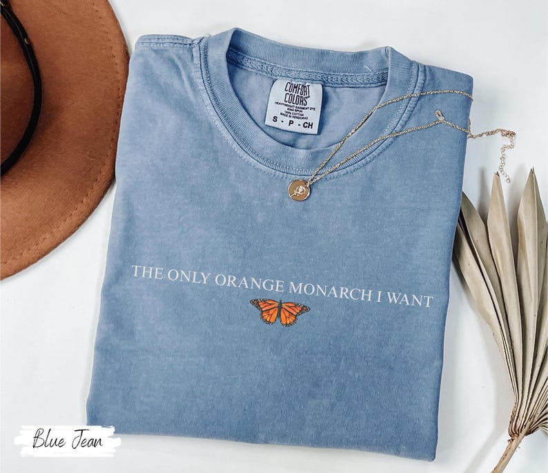 K&ouml;nnte beinhalten: Blaues Jeans-T-Shirt mit dem Text "THE ONLY ORANGE MONARCH I WANT" und einer Schmetterlingsgrafik. Eine goldene Halskette mit Anh&auml;nger liegt auf dem Shirt. Ein brauner Hut und getrocknete Palmbl&auml;tter sind im Hintergrund.