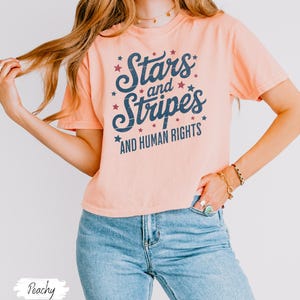 Puede incluir: Camiseta color melocot&oacute;n con el texto "Stars and Stripes AND HUMAN RIGHTS" en letras azul marino y detalles de estrellas rojas. La camiseta se combina con vaqueros azules claros.