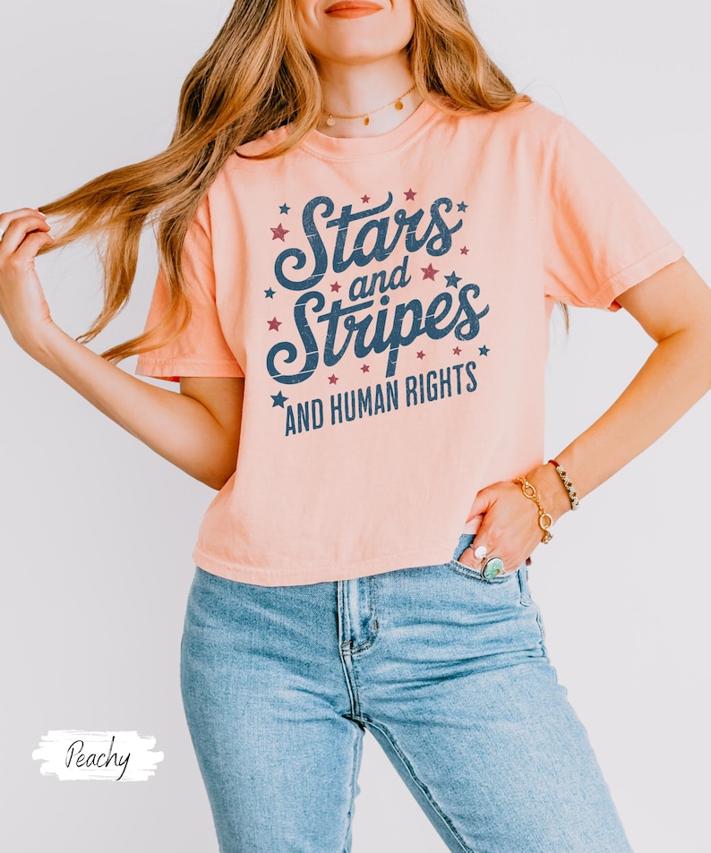 Puede incluir: Camiseta color melocot&oacute;n con el texto "Stars and Stripes AND HUMAN RIGHTS" en letras azul marino y detalles de estrellas rojas. La camiseta se combina con vaqueros azules claros.