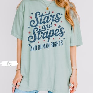 Puede incluir: Camiseta verde claro con el texto "Stars and Stripes AND HUMAN RIGHTS" en azul marino y rojo. El dise&ntilde;o incluye estrellas. La camiseta tiene un aspecto vintage y es adecuada para un estilo casual.
