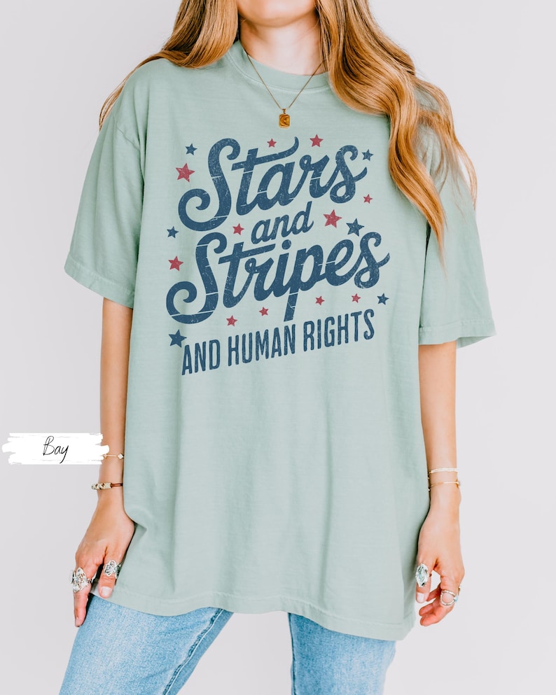 Puede incluir: Camiseta verde claro con el texto "Stars and Stripes AND HUMAN RIGHTS" en azul marino y rojo. El dise&ntilde;o incluye estrellas. La camiseta tiene un aspecto vintage y es adecuada para un estilo casual.