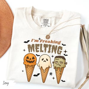 May include: Ivory-colored t-shirt with the text "I'm Freaking Melting" and Halloween-themed ice cream cone graphics. The cones feature a jack-o'-lantern, a ghost, and Frankenstein's monster. A gold necklace and a hat are also visible.