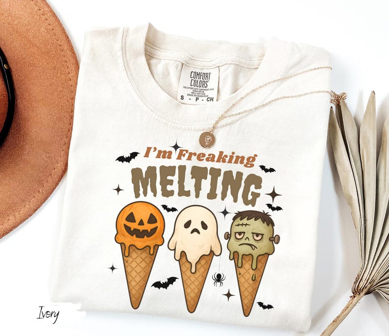 May include: Ivory-colored t-shirt with the text "I'm Freaking Melting" and Halloween-themed ice cream cone graphics. The cones feature a jack-o'-lantern, a ghost, and Frankenstein's monster. A gold necklace and a hat are also visible.
